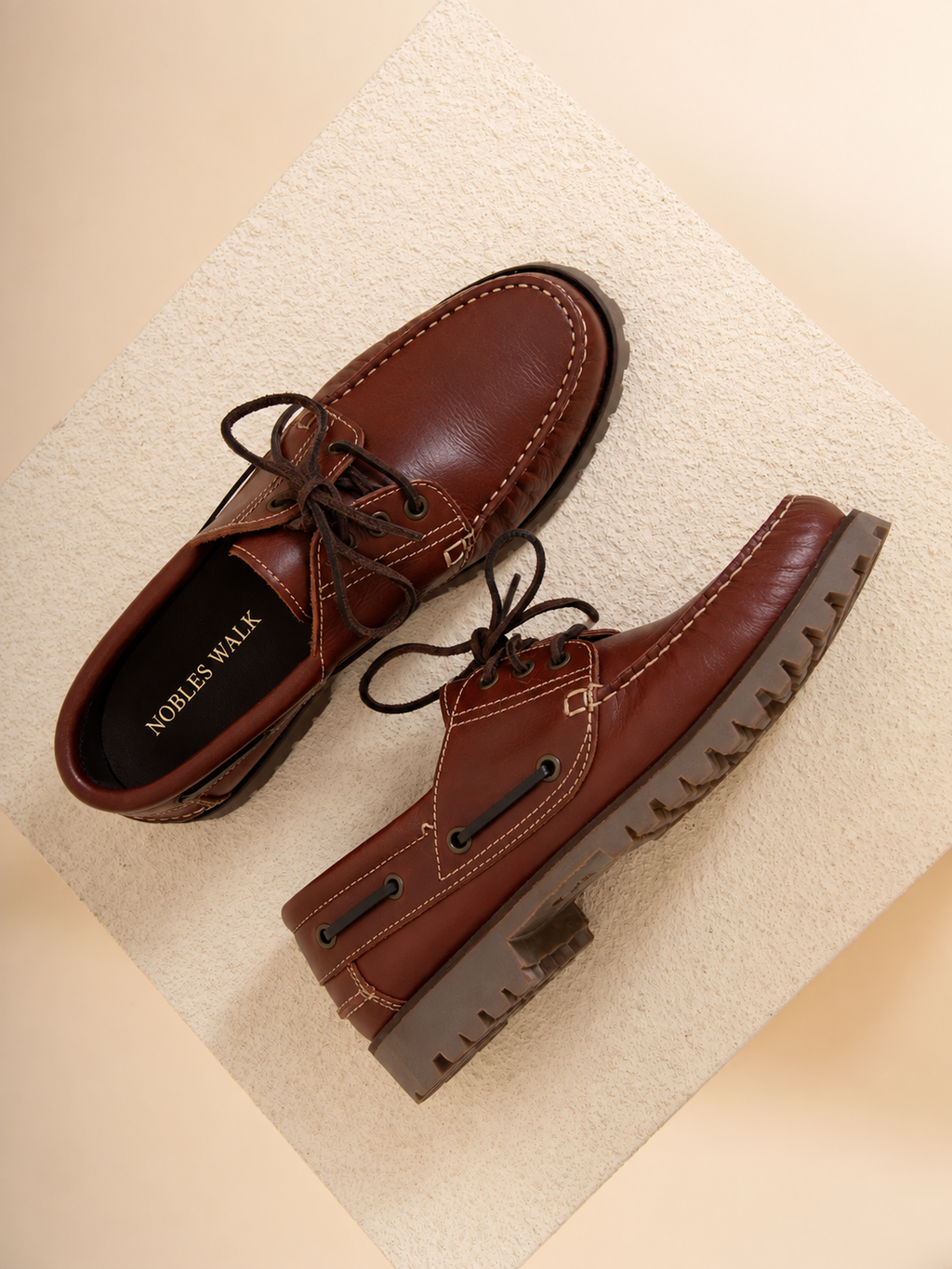 A pair of brown leather boat-style shoes with dark laces and rugged lug soles, displayed on a textured beige surface. The brand name 'NOBLES WALK' is visible on the insole of one shoe.