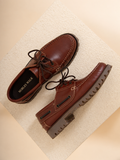 A pair of brown leather boat-style shoes with dark laces and rugged lug soles, displayed on a textured beige surface. The brand name 'NOBLES WALK' is visible on the insole of one shoe.