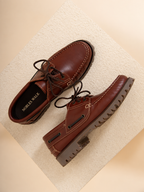 A pair of brown leather boat-style shoes with dark laces and rugged lug soles, displayed on a textured beige surface. The brand name 'NOBLES WALK' is visible on the insole of one shoe.